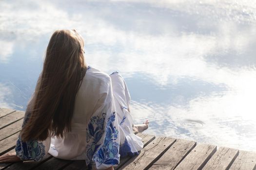 a girl in an embroidered Ukrainian shirt sits on the pier, the reflection of clouds in the water of the lake. On the shore of the sky. vyshyvanka day. freedom. patriot.