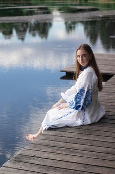 a girl in an embroidered Ukrainian shirt sits on the pier, the reflection of clouds in the water of the lake. On the shore of the sky. vyshyvanka day. freedom. patriot.