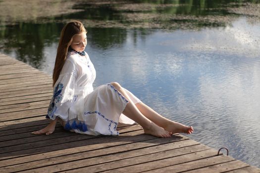 a girl in an embroidered Ukrainian shirt sits on the pier, the reflection of clouds in the water of the lake. On the shore of the sky. vyshyvanka day. freedom. patriot.