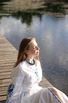 a girl in an embroidered Ukrainian shirt sits on the pier, the reflection of clouds in the water of the lake. On the shore of the sky. vyshyvanka day. freedom. patriot.