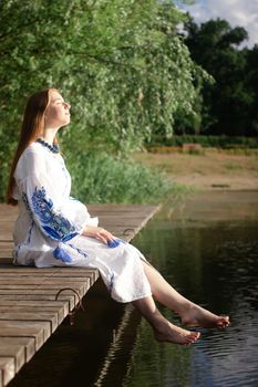 Girl in embroidered national Ukrainian costume on a pier on the shore of the lake. Independence day of ukraine, constitution, vyshyvanka day. young woman in blue dress outdoors.