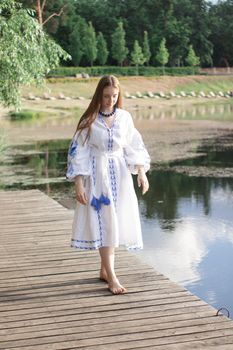 Girl in embroidered national Ukrainian costume on a pier on the shore of the lake. Independence day of ukraine, constitution, vyshyvanka day. young woman in blue dress outdoors.