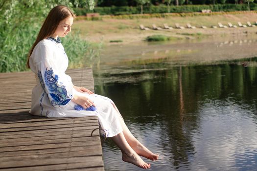 a girl in an embroidered Ukrainian shirt sits on the pier, the reflection of clouds in the water of the lake. On the shore of the sky. vyshyvanka day. freedom. patriot.