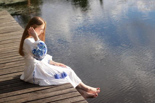 Girl in embroidered national Ukrainian costume on a pier on the shore of the lake. Independence day of ukraine, constitution, vyshyvanka day. young woman in blue dress outdoors.