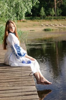 a girl in an embroidered Ukrainian shirt sits on the pier, the reflection of clouds in the water of the lake. On the shore of the sky. vyshyvanka day. freedom. patriot.