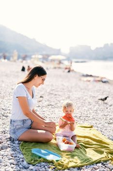 Smiling mom looking at baby girl eating watermelon on the beach. High quality photo