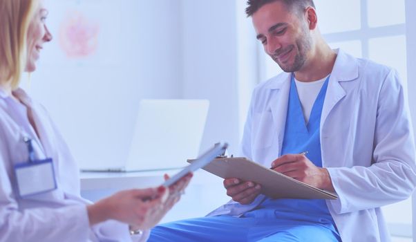 Handsome doctor is talking with young female doctor and making notes while sitting in his office