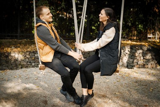 Young cute couple on a date looks at each other while sitting on a swing outdoor.