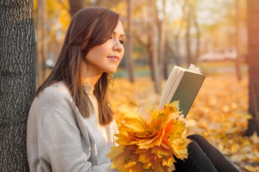 A young woman reads a book near a tree in an autumn park. The concept of self-development.