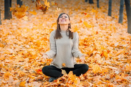 Smiling young woman sitting on the leaves having fun in autumn park throws up fallen leaves.