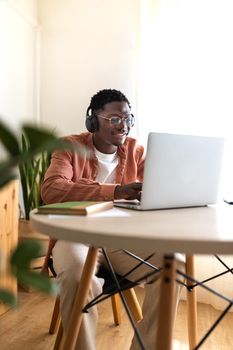 Vertical portrait of young African American man wearing headphones working, studying at home using laptop. Copy space. E-learning and technology concept.
