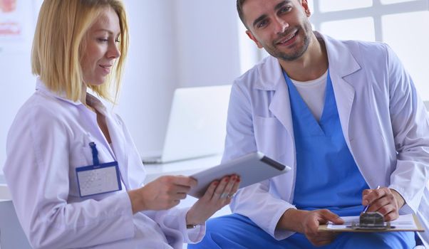 Handsome doctor is talking with young female doctor and making notes while sitting in his office