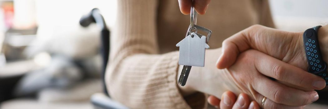 Close-up of realtor give keys from new accommodation to woman in wheelchair, kind handshake in agreement. Disability, government help, real estate concept