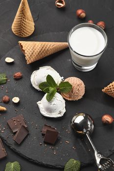 Top view of fantastic creamy and chocolate ice cream decorated with mint, a glass of milk, metal scoop, waffle cones with chocolate and hazelnuts nearby, served on a stone slate over a black background. Close-up.