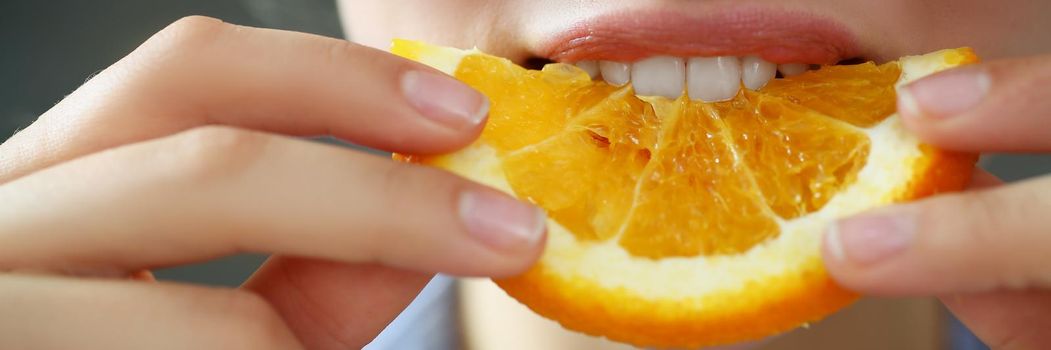 Close-up of woman hold in hand slice of chopped orange eats for breakfast with mouth in kitchen. Healthy eating, diet, juicy, fruit, summer harvest concept