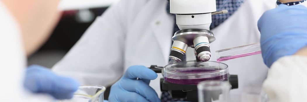 Close-up of female scientist explore purple sample under microscope in laboratory. Male chemist add liquid from flask. Lab, science, investigation concept