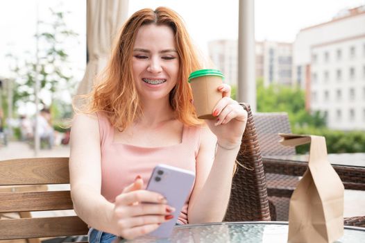 Young red-haired woman with braces on her teeth drinks coffee on a summer veranda