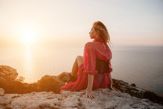 Side view a Young beautiful sensual woman in a red long dress posing on a volcanic rock high above the sea during sunset. Girl on the nature on blue sky background. Fashion photo