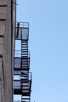 Silhouette of a fire escape on a high-rise building against a blue sky with clouds. Some of the stairs are broken. There is free space for text