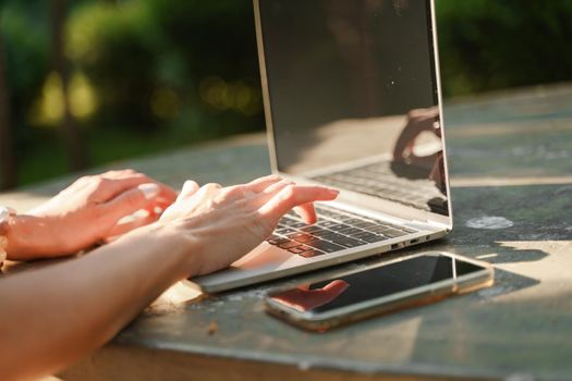 a beautiful mature woman in hat works on a computer at a white table in nature and spends her day productively.