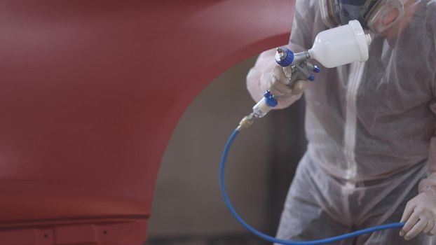 Red car in a paint chamber during repair work. Auto painting worker. Professional car painter is painting a body work. Male technician is air-painting car's door.
