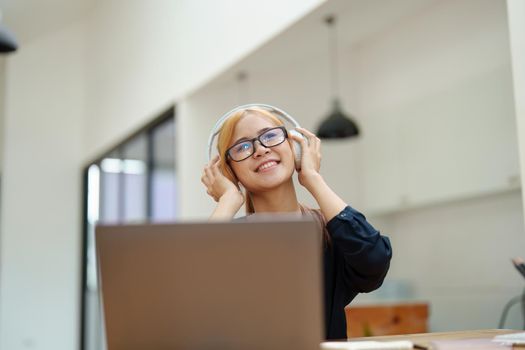 A portrait of a young Asian woman with blonde hair wearing over-ear headphones listening to music to relax while taking a break from boring day activities.