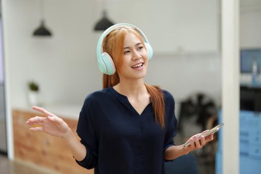 A portrait of a young Asian woman with blonde hair wearing over-ear headphones listening to music to relax while taking a break from boring day activities.