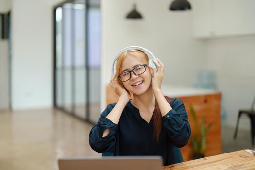 A portrait of a young Asian woman with blonde hair wearing over-ear headphones listening to music to relax while taking a break from boring day activities.