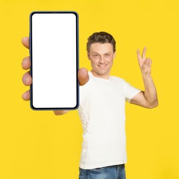 Gesturing V or victory handsome man holding huge smartphone with white screen wearing white t-shirt and jeans isolated on yellow background. Mobile app advertisement, great offer.