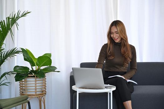 Cheerful asian woman surfing internet with computer laptop in living room.