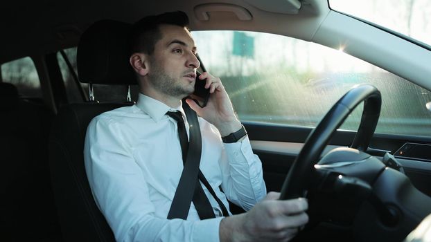 Handsome young man driver riding car and speaking on phone, having conversation using smartphone, talking while driving. Lifestyle, road, car concept.