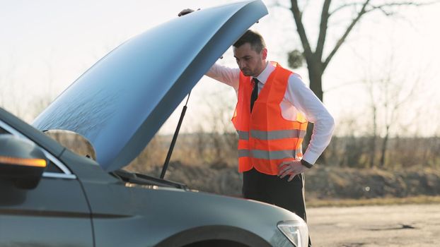 Disappointed man in formal outfit opening bonnet of broke down car to check engine. Sad businessman standing near car opened the hood. Car accident on the road. Emergency stop sign