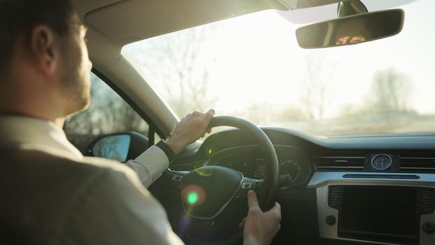 Man driving car towards bright scenery sun pleasure shining over the road. Male Hand on steering wheel. Man Driving a Car at Sunset. Elegant male driver traveling by automobile.