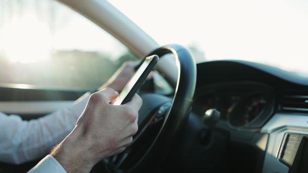 Close-up hands of unrecognizable man typing online message using mobile phone while sitting at car. Closeup view of businessman using phone in auto. Accident insurance. Chatting driver.