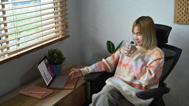 Casual young woman drinking coffee and browsing internet with laptop at home.