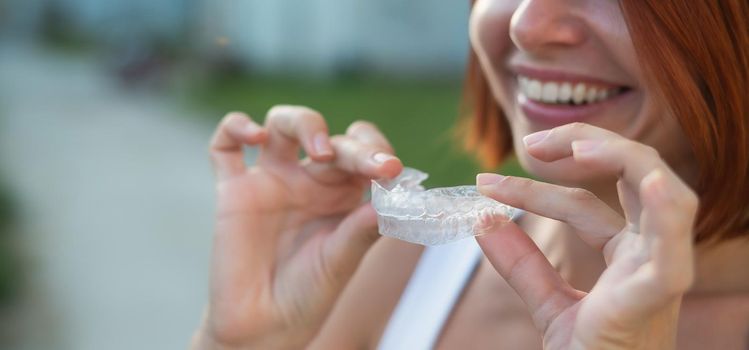 Red-haired Caucasian woman holding transparent mouthguards for bite correction outdoors. A girl with a beautiful snow-white smile uses silicone braces.