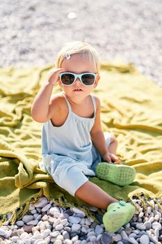 Little girl adjusts her sunglasses sitting on a blanket on the beach. High quality photo