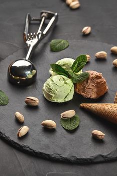 Delicious chocolate and pistachio ice cream decorated with mint, classic waffle cones with scattered nuts are nearby, served with a metal scoop on a stone slate over a black background. Close-up.