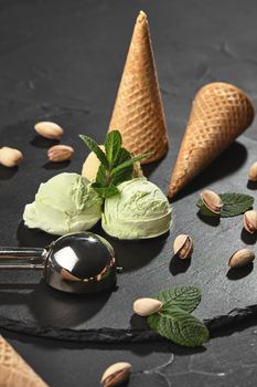Cold pistachio ice cream decorated with mint, waffle cones with scattered pistachios are nearby, served with a glossy scoop on a stone slate over a black background. Close-up.