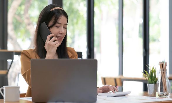 Beautiful Asian businesswoman analyzes charts using laptop calculator and talking the phone at the office..