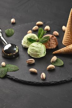 Gourmet chocolate and pistachio ice cream decorated with mint, classic waffle cones with scattered nuts are nearby, served with a metal scoop on a stone slate over a black background. Close-up.