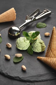 Enjoyable pistachio ice cream decorated with mint, waffle cones with scattered pistachios are nearby, served with a glossy scoop on a stone slate over a black background. Close-up.