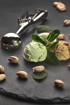 Close-up shot of a tasty chocolate and pistachio ice cream decorated with mint, scattered pistachios are nearby, served with a glossy scoop on a dark slate over a black background.