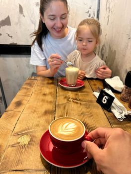 Mom treats a little girl to a latte from a spoon at the table. High quality photo
