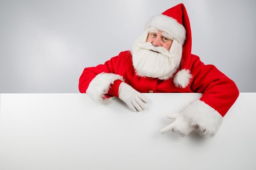 Santa Claus peeks out from behind a white background and points to an empty space with his finger. Merry Christmas