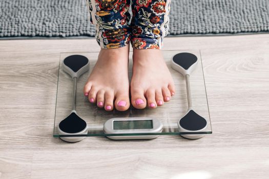 Legs of a girl standing on scale to measure weight. Caucasian female bare feet with weight scale at home. Person checking the weight on the scale. Dieting, control and measuring