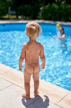 Small naked baby stands by the pool with turquoise water. Back view. High quality photo