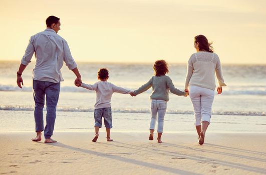 Rear view shot of a carefree family holding hands while walking on the beach at sunset. Mixed race parents and their two kids spending time together by the sea enjoying summer vacation.