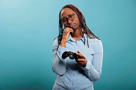 African american woman got upset because of lost online competitive videogame match. Sad young adult lady with modern gaming console controller standing on blue background.