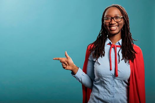 Joyful and confident superhero woman wearing red cape while pointing fingers to left on blue background. Selfless and strong young adult justice defender person posing at camera. Studio shot.